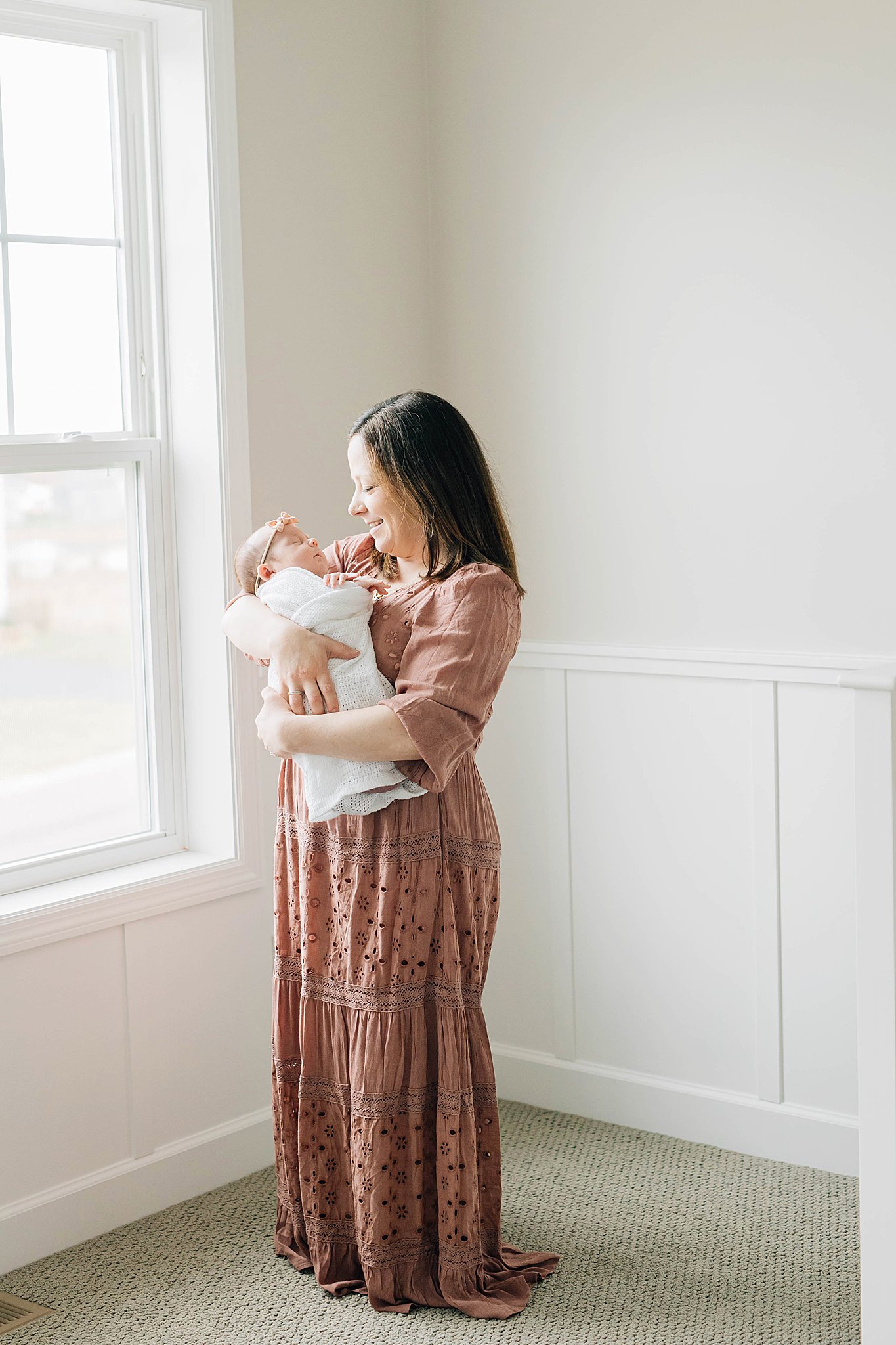 Mom and newborn by window, with mom wearing long dress for outfit for newborn photos. 