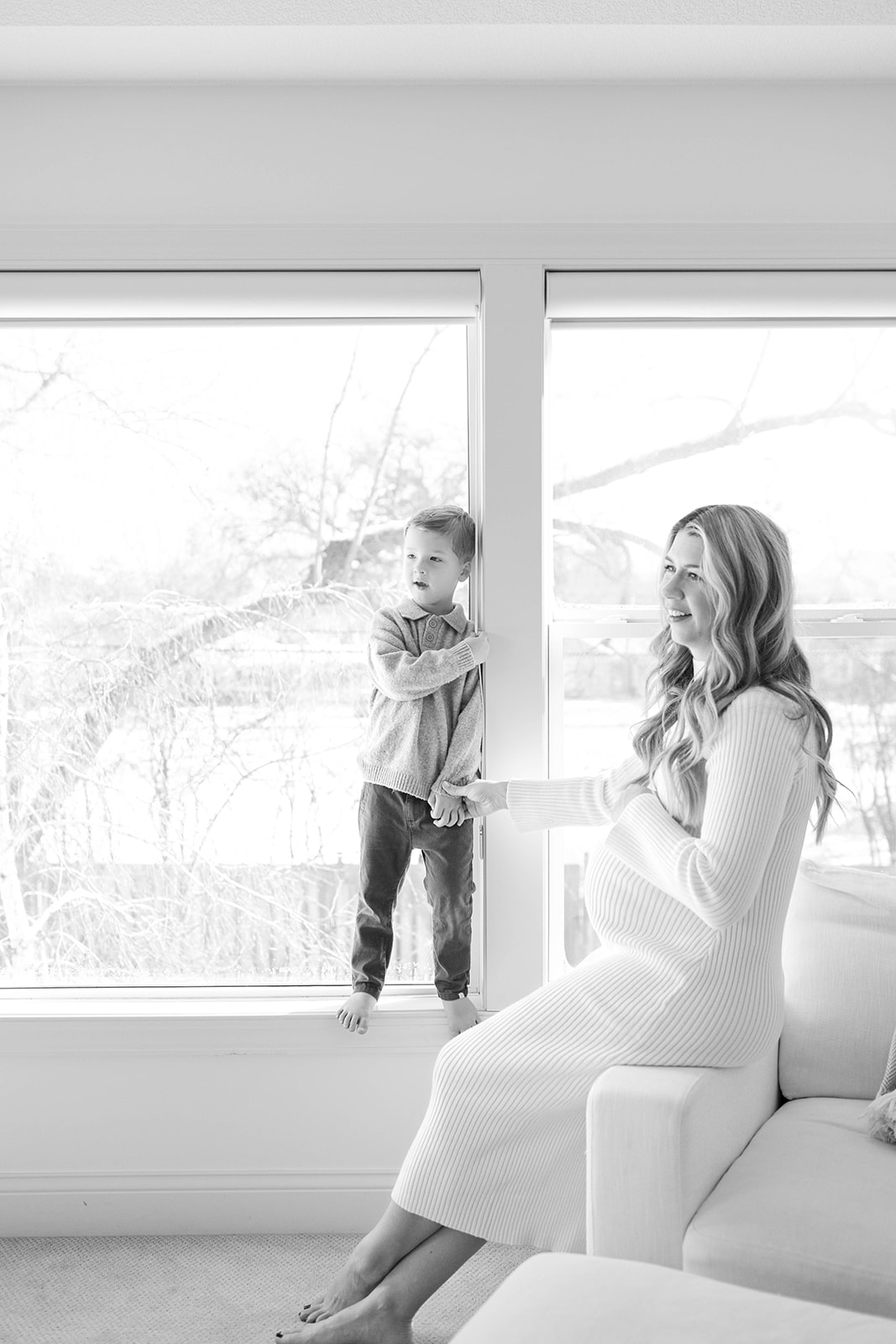 Toddler Boy Stands on Windowsill, Holding Moms Hand, During Family Photos